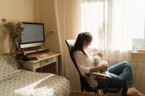 Adulto mamma in abbigliamento casual succhiare affascinante bambino mentre seduto in camera luce casa — Foto stock
