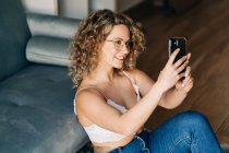 From above of self assured young female millennial with curly blond hair in leather bra an jeans sitting on floor near sofa and taking selfie on smartphone — Foto stock