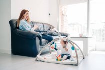 Окаменелая молодая мама в повседневной одежде, просматривающая смартфон и нетбук сидя на диване рядом с обожаемым малышом, играющим с игрушками на полу в гостиной — стоковое фото