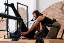 Flexible female stretching legs with help of personal instructor while doing exercises on pilates reformer — Stock Photo