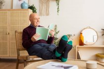 Sonriente hombre maduro sentado en silla de madera y libro de lectura mientras disfruta de fin de semana en acogedora sala de estar - foto de stock