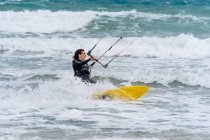 Atleta feminina ativa no kiteboard segurando barra de controle enquanto pratica kitesurf e olhando para o oceano espumoso — Fotografia de Stock