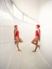 Side view of young female athletes in same swimwear with raised legs standing on tiled floor in passage — Stock Photo