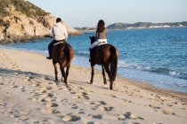 Back view of unrecognizable young woman with boyfriend riding purebred stallions on sandy shore against wavy ocean under blue sky — Stock Photo