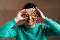 Cheerful male broker in green wear covering eyes with metal crypto coins on brown background — Stock Photo