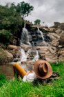 Anonimo viaggiatore maschio in cappello rilassante sul lungolago lussureggiante e godendo di una rapida vista a cascata nella natura estiva — Foto stock