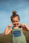 Очаровательная маленькая девочка в комбинезоне, стоящая с пальцами на лице на лугу и смотрящая вниз — стоковое фото