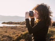 Боковой вид сфокусированной молодой кудрявой этнической женщины-путешественницы, фотографирующей на фотокамеру, стоя у моря в солнечный вечер — стоковое фото
