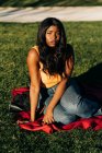 Femme afro-américaine insouciante en tenue tendance assise sur un plaid sur la pelouse dans le parc et regardant la caméra — Photo de stock