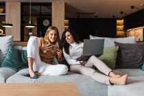 Couple of cheerful homosexual women chilling on couch and watching video on smartphone and working on laptop while entertaining together at weekend — Stock Photo