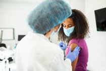 Specialista in uniforme protettiva, guanti in lattice e maschera facciale che vaccina la paziente afroamericana in clinica durante l'epidemia di coronavirus — Foto stock