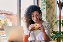 Contenido étnico femenino remoto trabajador con sabroso sándwich de bagel vegetariano contra netbook y bebida refrescante en el restaurante - foto de stock