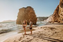 Полнотелый мужчина обнимает молодую привлекательную девушку в купальниках, расслабляясь вместе на песчаном побережье у грубых скал в солнечный день в Фириплака Милош — стоковое фото