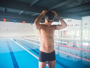 Sportsman in swimming trunks and cap with hands behind head preparing for training against pool with lanes in building — Stock Photo