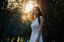 Вид збоку спокійна жінка в білій сукні, що стоїть на стовбурі дерева в темних лісах у спокійному сонячному світлі — стокове фото