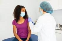 Specialista in uniforme protettiva, guanti in lattice e maschera per il viso che vaccina la paziente afroamericana in clinica durante l'epidemia di coronavirus — Foto stock