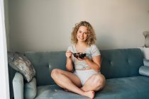 Full length cheerful female in domestic wear enjoying yummy food in bowl while sitting on comfortable sofa with legs crossed and looking at camera with smile — Fotografia de Stock