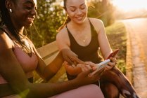 Весела багаторасова жінка-спортсменка в активному одязі сидить на лавці в парку і використовує мобільні телефони разом після тренування на заході сонця — стокове фото