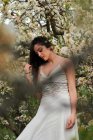 Junge Frau mit tätowiertem Arm trägt weißes Kleid und steht in Blumen des Baumes und blickt nach unten — Stockfoto
