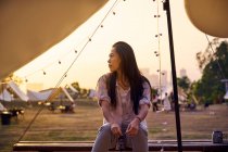 Belle femme asiatique ethnique assise à table tout en ayant un moment de détente dans la zone de camping pendant les vacances pendant le coucher du soleil regardant loin — Photo de stock