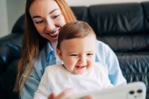 Glückliche junge Mutter und entzückendes lächelndes Baby, das lustige Videos auf dem Handy anschaut, während es zu Hause auf dem gemütlichen Sofa sitzt — Stockfoto