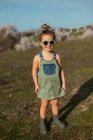 Content petite fille en salopette et lunettes de soleil debout dans la prairie et profiter de l'été par une journée ensoleillée à la campagne — Photo de stock