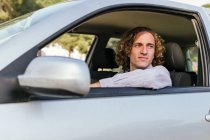 Happy young haired male looking away through open window of car while sitting at driver seat — Fotografia de Stock