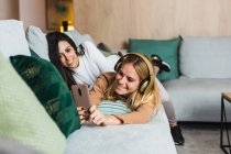 Un par de mujeres lesbianas tumbadas en el sofá y tomando fotos en el teléfono inteligente mientras se relajan juntas en la sala de estar el fin de semana - foto de stock