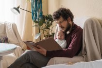 Barbuto papà con adorabile bambino lettura libro mentre seduto con le gambe incrociate in poltrona in casa — Foto stock