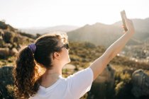 Back view of happy young female traveler with long dark hair in casual clothes and sunglasses taking selfie on mobile phone during hiking trip in mountainous terrain — Stock Photo