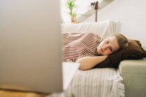 Jeune femme épuisée couchée sur le canapé et regardant un film sur netbook dans le salon à la maison — Photo de stock