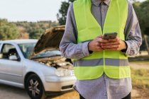 Crop unrecognizable male driver in green safety vest using mobile phone and asking for help after car accident on country road — Foto stock