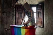 Невпізнавана людина в срібному костюмі і коробці на голові прасує прапор ЛГБТК, стоячи в покинутій кімнаті — стокове фото