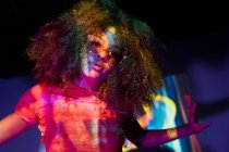 Portrait de la jeune femme afro-américaine attrayante dans des lunettes de soleil élégantes regardant la caméra tout en se tenant debout dans des néons — Photo de stock