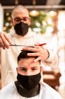Male hairstylist in eyeglasses making haircut to adult client in hairdressing salon during COVID 19 pandemic — Photo de stock