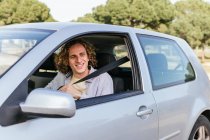 Happy young haired male looking away through open window of car while sitting at driver seat — Foto stock