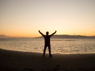 Back view silhouette of male traveler with arms raised enjoying freedom while standing on seashore at sunset time — Stock Photo