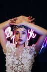 Charming tender young bride in white lace gown and luxurious floral wreath and earrings looking at camera against black background with neon lights — Fotografia de Stock