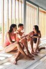 Cheerful sportsman with content female athletes in swimsuits sitting with crossed legs on tiled floor after workout — Stock Photo