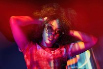 Attraktive junge Afroamerikanerin mit stylischer Sonnenbrille, die lockiges Haar berührt und im Neonlicht in die Kamera schaut — Stockfoto