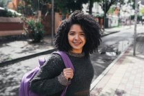 Side view of delighted ethnic female student with Afro hairstyle and backpack standing on street on sunny day and looking at camera — Foto stock