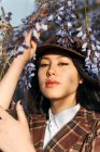 Young gentle ethnic female in checkered wear with beret touching blossoming plant while looking at camera on street — Stock Photo