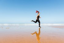 Vista laterale del jogger femminile attivo con tatuaggi che corrono sulla riva dell'oceano mentre riflettono in acqua durante l'allenamento — Foto stock