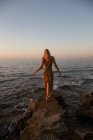 Visão traseira da jovem mulher andando na costa contra o mar azul acenando ao pôr do sol — Fotografia de Stock