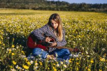 Junge Hipster-Frau sitzt auf einer Wiese in der Natur und schreibt Lieder auf einem Notizbuch und spielt in der Sommersonne Gitarre. — Stockfoto