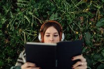 Top view of young attentive female in modern headset reading textbook while lying on meadow in summer - foto de stock