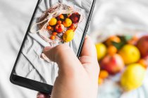 Overhead view of crop unrecognizable person touching screen on mobile phone while taking photo of fruits in zero waste bag - foto de stock