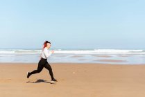 Vista lateral da jogger fêmea ativa correndo na costa oceânica durante o treinamento no verão — Fotografia de Stock