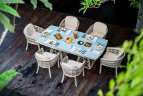 Сверху удобное плетеное кресло с мягкими креслами и подушками, расположенными рядом со столом, подаваемым с разнообразной аппетитной выпечкой и фруктами во время завтрака в тропическом курорте — стоковое фото