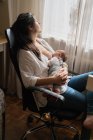 Maman adulte en tenue décontractée allaitant charmant petit enfant assis dans la pièce de la maison légère — Photo de stock